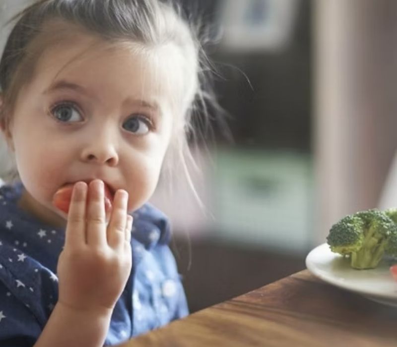 A toddler tasting food at the table with broccoli on a plate nearby
